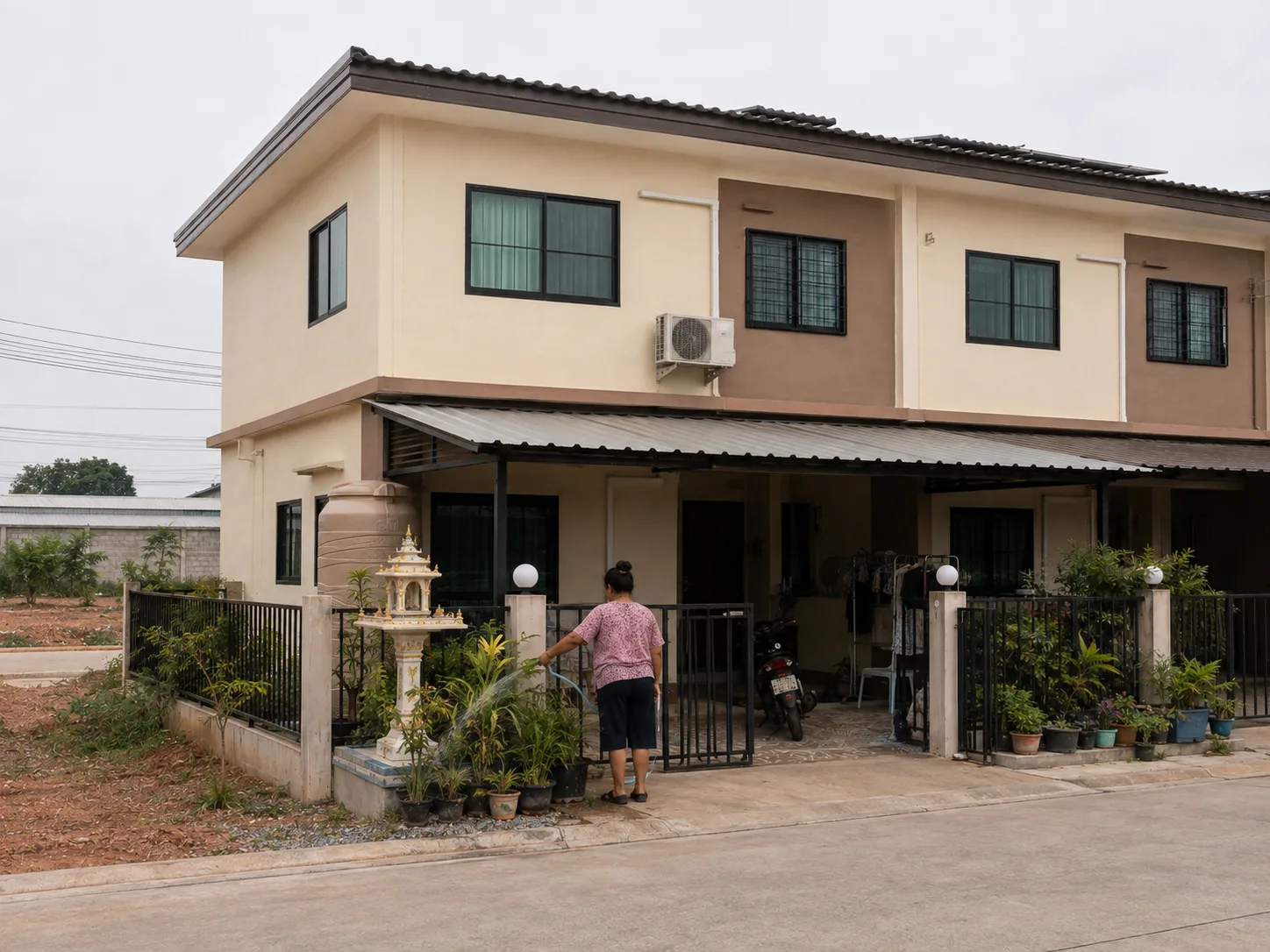 ภายนอกบ้านทาวน์เฮาส์ของรากฐาน ผนังสีครีม ที่จอดรถมีหลังคา แนวหน้าบ้านเล็ก ๆ ใต้ท้องฟ้าครึ้ม